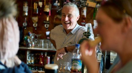 Een oudere man met een vrolijke uitdrukking staat zelfverzekerd achter een bar, die doet denken aan een gezellige pub op een rondreis Ierland. Omringd door flessen en barparafernalia weerspiegelt de warme, gezellige setting de uitnodigende charme van een onvergetelijke vakantie Ierland. | Echt Ierland Vakanties