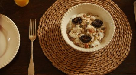 Een witte kom gevuld met ontbijtgranen, yoghurt, pruimen en zaden staat op een geweven placemat. Links roept een wit bord met een gevouwen rode servet en hoog gras herinneringen op aan een urlaub Irland-avontuur. Er ligt een vork naast. Rechts liggen een mes, lepel en klein bordje. Op de achtergrond staat een glas sinaasappelsap. | Echt Ierland Vakanties