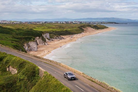 Een schilderachtige kustweg, perfect voor een vakantie Ierland avontuur, met een auto die erlangs rijdt. Beneden is een zandstrand met verspreide mensen. De kustlijn heeft groene kliffen en een kalme oceaan onder een gedeeltelijk bewolkte lucht, terwijl verre heuvels en een charmant stadje de achtergrond vormen. | Echt Ierland Vakanties