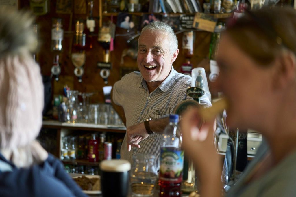 Een man in een licht shirt lacht warm achter een bar tijdens een stop in Ierland, in gesprek met twee gasten. De voorgrond toont een wazig persoon die aan een drankje nipt. De toonbank bevat een pint stout en fruitdrank terwijl planken, flessen en decor de achtergrond vullen. | Echt Ierland Vakanties