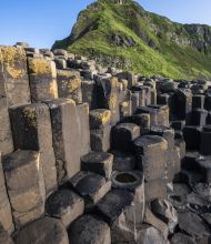 De afbeelding toont de Giant's Causeway, een must-see op *rondreis Ierland*, met hexagonale basaltkolommen. Ongelijke verweerde kolommen met geel korstmos staan op de voorgrond, terwijl een grasheuvel onder een helderblauwe lucht dit iconische Ierse tafereel compleet maakt. | Echt Ierland Vakanties