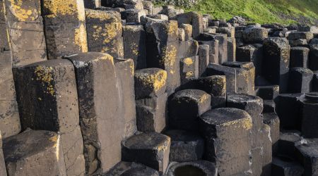 De afbeelding toont de Giant's Causeway, een must-see op *rondreis Ierland*, met hexagonale basaltkolommen. Ongelijke verweerde kolommen met geel korstmos staan op de voorgrond, terwijl een grasheuvel onder een helderblauwe lucht dit iconische Ierse tafereel compleet maakt. | Echt Ierland Vakanties