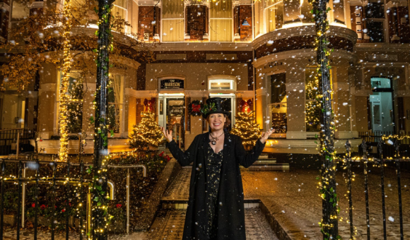 Een persoon in een zwarte jas en hoge hoed staat vrolijk in de vallende sneeuw, armen omhoog. Ze staan voor een versierd gebouw versierd met warme lichten, kransen en kerstbomen. Deze besneeuwde scène roept de feestelijke sfeer op van een winternacht, net als een charmante Ierland vakantie-ervaring. | Echt Ierland Vakanties