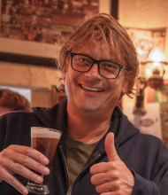 Lachende man met bril en warrig haar houdt een drankje in zijn rechterhand en steekt zijn duim omhoog met zijn linkerhand, wat de geest van "urlaub Irland" belichaamt. Hij draagt een marineblauw jasje. Warme binnenverlichting met ingelijste foto's aan de muur op de achtergrond. | Echt Ierland Vakanties