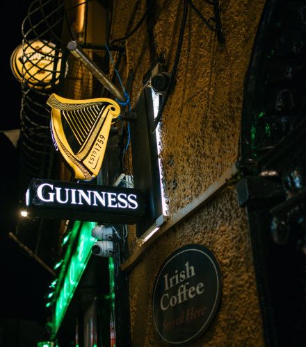's Avonds hangt er een gloeiend Guinness-bord in de vorm van een harp buiten een pub – een welkome aanblik tijdens elke herfstrondreis door Ierland. Daaronder hangt een rond bord met de tekst "Irish Coffee Served Here". Groene lampjes en slingers dragen bij aan de levendige sfeer tegen de donkere hemel. | Echt Ierland Vakanties