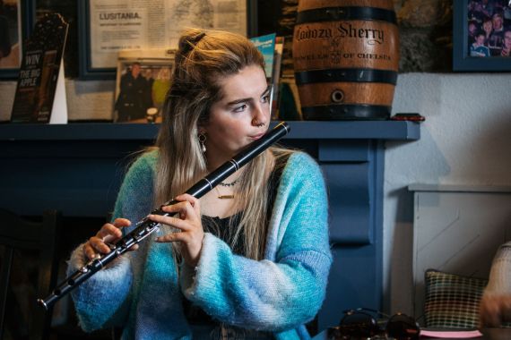 Een vrouw met lang blond haar, gekleed in een blauw vest, bespeelt een zwarte fluit in een gezellige pub tijdens een herfstrondreis door Ierland. Zonlicht accentueert haar gelaatstrekken, terwijl ingelijste foto's en een groot sherryvat de perfecte achtergrond vormen. | Echt Ierland Vakanties