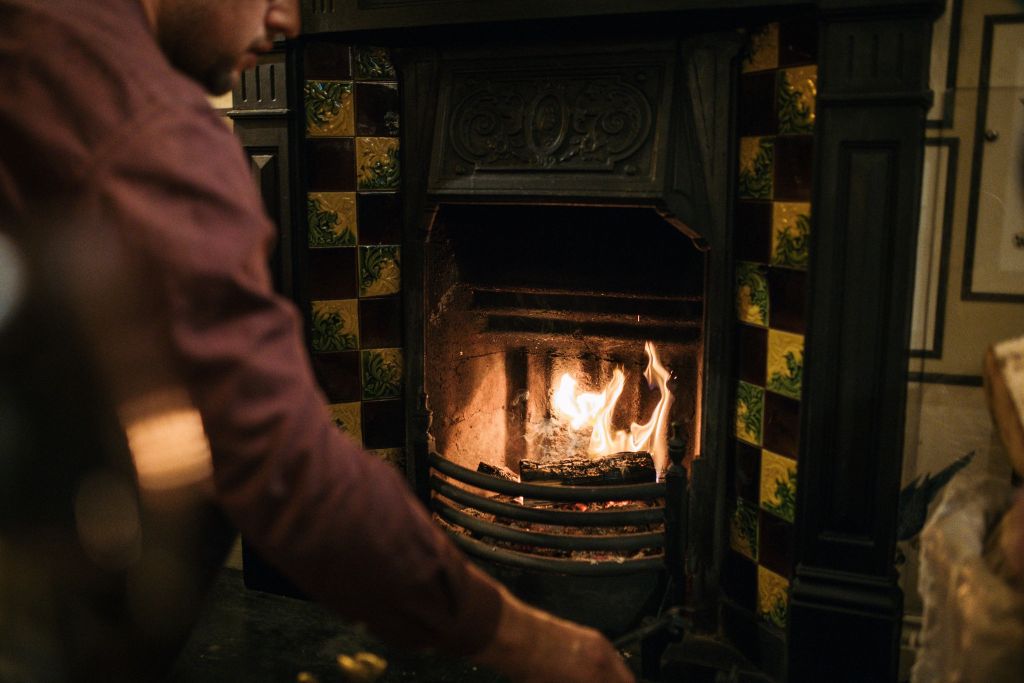 Een persoon in een bordeauxrood overhemd houdt een klein vuurtje aan in een sierlijke, donkergekleurde open haard met decoratieve geel-groene tegels. De gezellige, warm verlichte kamer roept de uitnodigende sfeer op van een herfstrondreis door Ierland. | Echt Ierland Vakanties