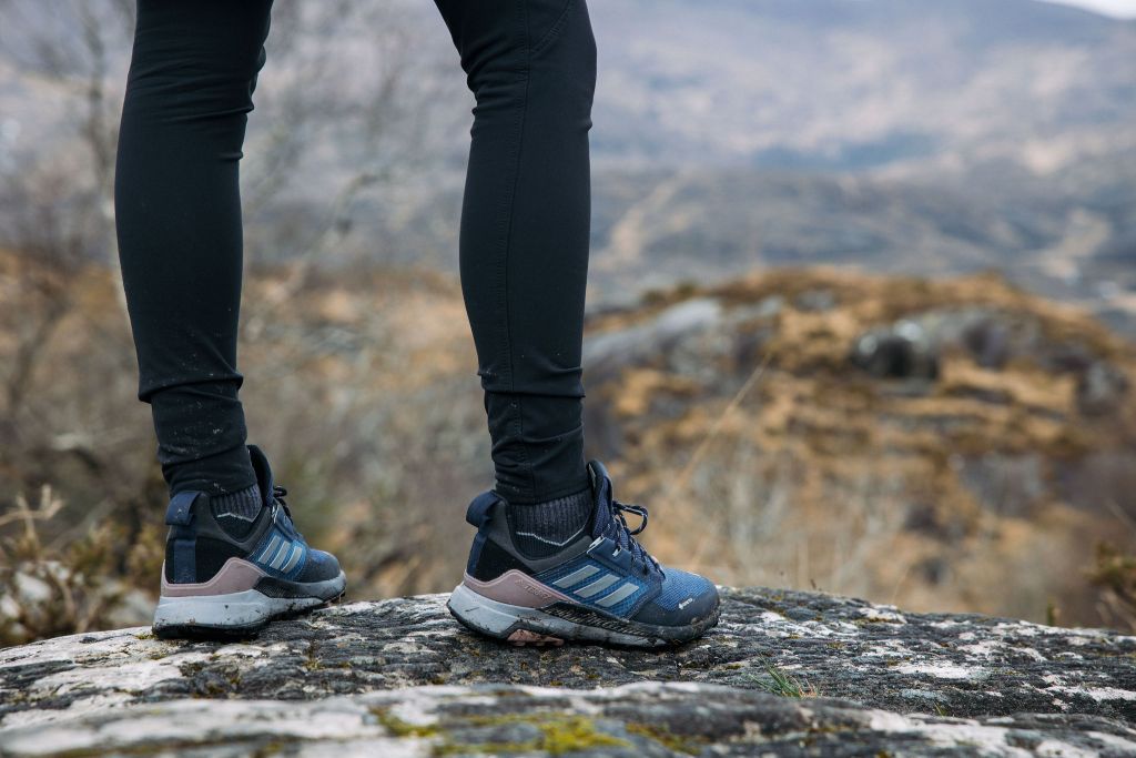 Een persoon in een zwarte legging en blauwe Adidas-wandelschoenen staat op een met mos begroeide rots. Het wazige, ruige landschap doet denken aan een herfstrondreis door Ierland, met bruine heuvels en rotsachtig terrein als perfecte achtergrond voor een buitenavontuur. | Echt Ierland Vakanties