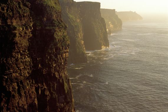 Steile kliffen bedekt met groene grasvelden rijzen dramatisch uit boven de oceaan bij zonsondergang – een iconisch gezicht tijdens een herfstrondreis door Ierland. Zacht licht baadt in het landschap terwijl een kalme zee en een wazige lucht een vredige, majestueuze sfeer creëren. | Echt Ierland Vakanties