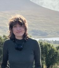 Een persoon met golvend haar in een donkergroen shirt met lange mouwen staat buiten, wat wijst op een inspirerende rondreis Ierland. Met een verrekijker om hun nek bewonderen ze een schilderachtig landschap met bergen, bomen en water onder een gedeeltelijk bewolkte lucht. Het licht creëert een serene ambiance. | Echt Ierland Vakanties