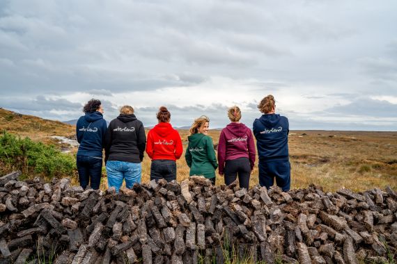 Vijf mensen met hun rug naar elkaar, met hoodies in zwart, rood, groen en blauw, elk geborduurd met tekst. Ze staan op een grasvlakte achter stapels turfstenen. De lucht is bewolkt en de horizon is ver weg: een perfect tafereel voor een vakantie in Ierland die de landelijke essentie van een Iers landschap vastlegt. | Echt Ierland Vakanties