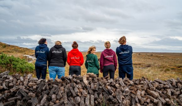 Vijf mensen met hun rug naar elkaar, met hoodies in zwart, rood, groen en blauw, elk geborduurd met tekst. Ze staan op een grasvlakte achter stapels turfstenen. De lucht is bewolkt en de horizon is ver weg: een perfect tafereel voor een vakantie in Ierland die de landelijke essentie van een Iers landschap vastlegt. | Echt Ierland Vakanties