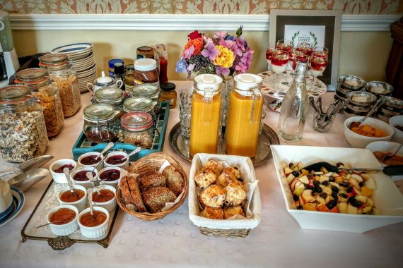 Een ontbijtbuffet bestaat uit ontbijtgranen, brood, kleine jampotjes, gebak en sinaasappelsapdispensers. Waterflessen en een schaal fruitsalade met een serveerlepel accentueren de spread. Kleine dessertglaasjes bevatten yoghurt of gelaagd fruit. Een bloemstuk voegt charme toe, wat de schoonheid van een vakantie in Ierland weerspiegelt. | Echt Ierland Vakanties