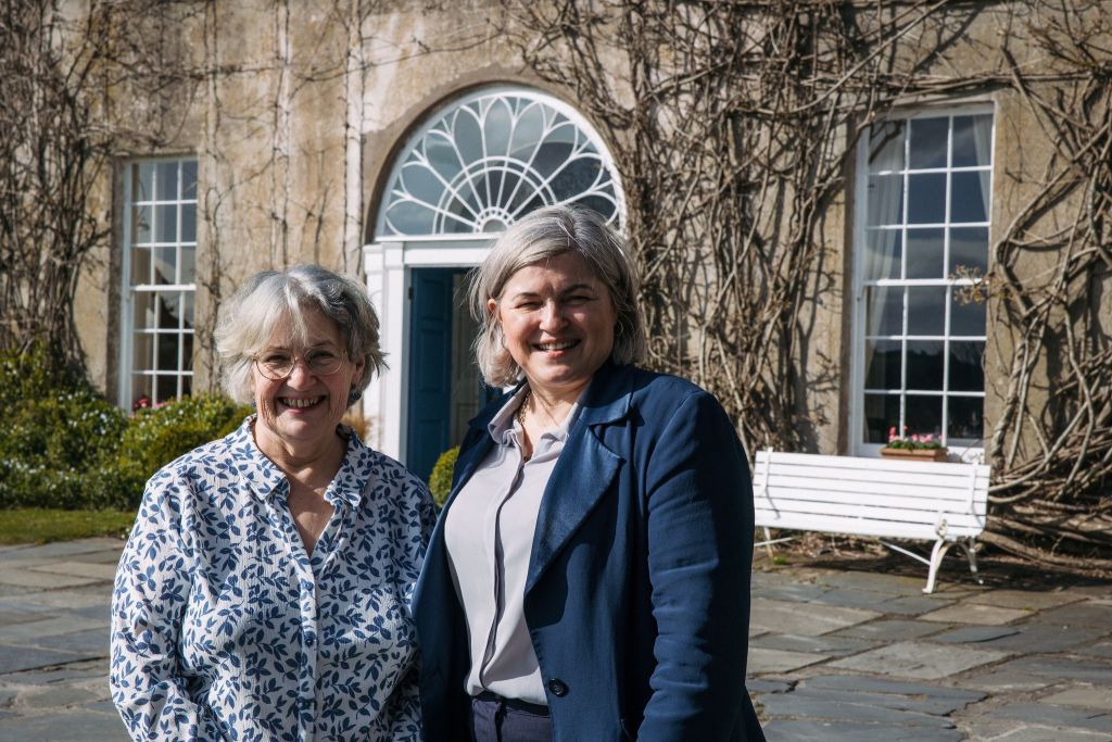 Twee vrouwen lachen voor een historisch gebouw met een stenen gevel en klimplanten. De een draagt een bril en een blauw gebloemd bloesje; de ander draagt een blauwe blazer en een grijs bloesje. Achter hen is een boograam en een witte bank. De zon schijnt fel. | Echt Ierland Vakanties