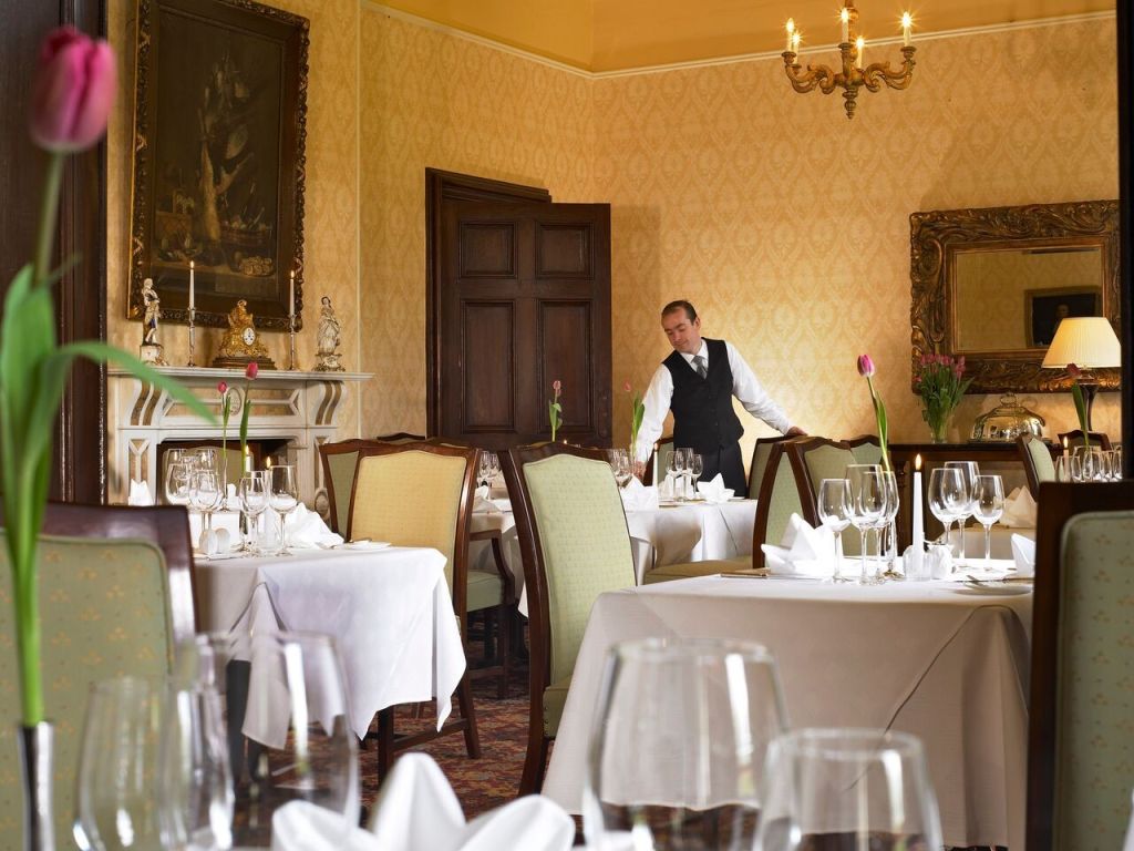 Een ober in een formeel restaurant met vintage decor, inclusief een kroonluchter, sierlijke spiegel, behang met patroon en een groot schilderij. Tafels zijn gedekt met witte tafellakens, wijnglazen en gevouwen servetten. Twee hoge tulpen in vazen voegen een vleugje kleur toe aan de elegante sfeer. | Echt Ierland Vakanties