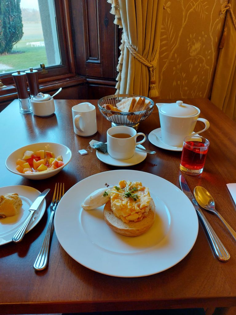 Een houten tafel voor het ontbijt bij een raam. Er staat een bord met een Engelse muffin met roerei en kruiden erop. Ernaast staan een fruitschaal, een kop zwarte koffie, een glas sap, boter en een theepot. Op de achtergrond is een tuin te zien door het raam. | Echt Ierland Vakanties