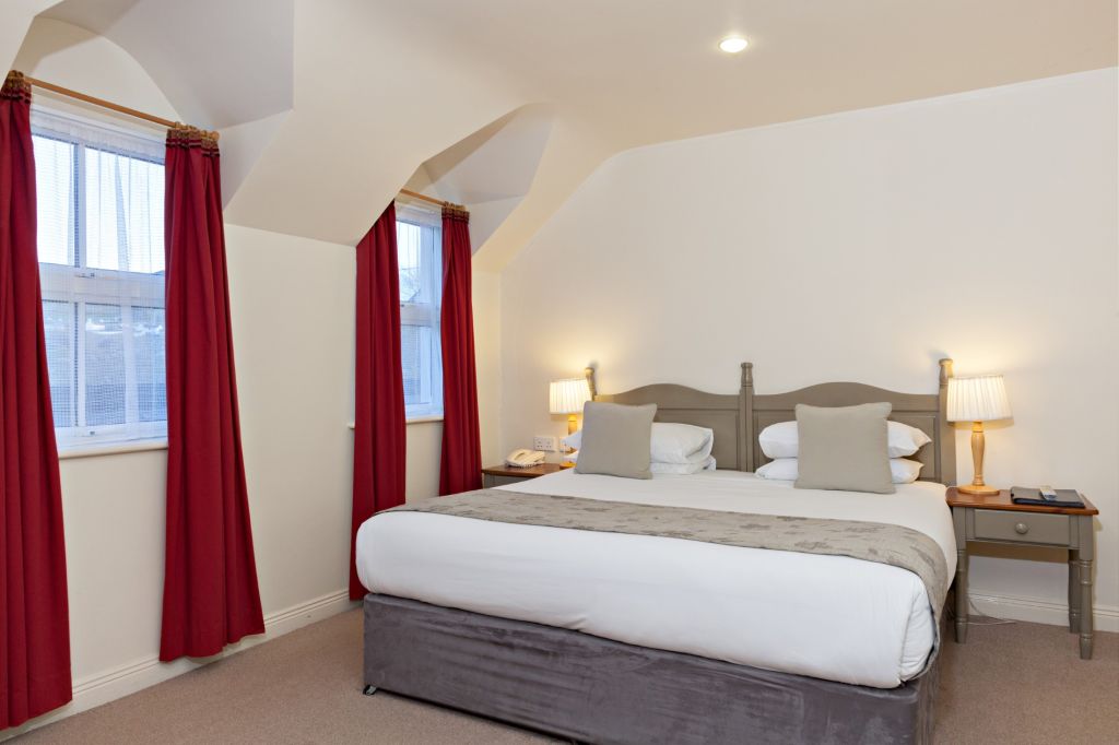 Een gezellige slaapkamer met een groot bed met wit linnen en een grijze sprei. Het bed heeft twee grijze kussens. Twee nachtkastjes met lampen staan naast het bed. De kamer heeft beige muren, rode gordijnen en twee ramen die natuurlijk licht binnenlaten. Het plafond is schuin, met één plafondlamp. | Echt Ierland Vakanties