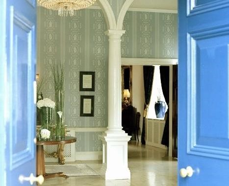 De afbeelding toont een elegante hotelfoyer met open blauwe dubbele deuren. Een grote kroonluchter hangt boven een patroonblauw tapijt, wat de charme van Dunbrody Country House oproept. Witte zuilen omlijsten een tafel versierd met planten, terwijl ingewikkeld behang de verfijnde ambiance versterkt. | Echt Ierland Vakanties