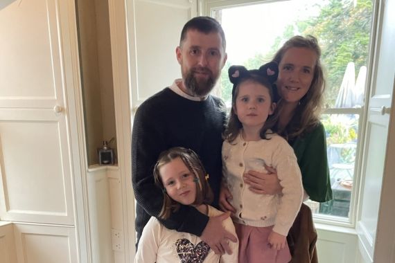 Een gezin van vier staat binnen bij een raam. Een man met een baard staat links en een vrouw met lang haar staat rechts. Twee jonge meisjes staan tussen hen in; een omhelst de vrouw. Bomen zijn zichtbaar buiten het raam, wat groen toevoegt aan de achtergrond. | Echt Ierland Vakanties