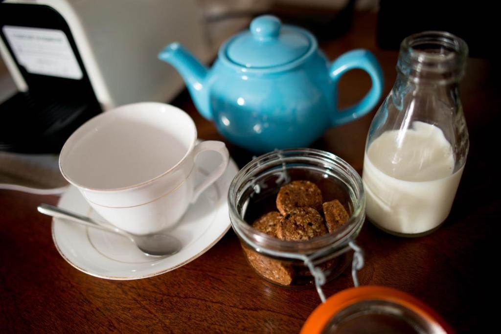 Een blauwe theepot, een witte theekop met een schotel en lepel, een glazen pot met koekjes en een klein flesje melk staan op een houten tafel. De theekop is leeg en de koekjes zijn zichtbaar door het open deksel van de pot. | Echt Ierland Vakanties