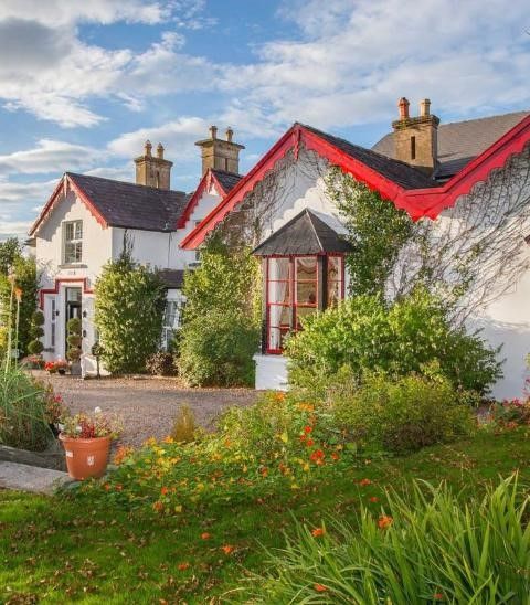Een charmant wit huisje met rode randen, genesteld in een weelderige tuin. Het huis heeft met klimop begroeide muren, verschillende ramen met rode kozijnen en een grindpad dat naar de ingang leidt. Levendige bloeiende planten omringen het gazon en een klassieke lantaarnpaal staat bij het pad onder een gedeeltelijk bewolkte lucht. | Echt Ierland Vakanties