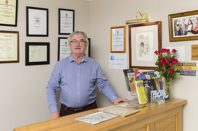 Een man met grijs haar, een bril en een blauw overhemd staat achter de houten receptie van Fanad House Bed and Breakfast. Brochures, een kaart en een vaas met rode rozen staan op de balie; Kilkenny-certificaten en onderscheidingen hangen aan de beige muur achter hem. | Echt Ierland Vakanties