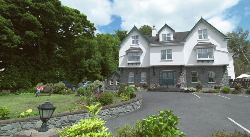 Het grote witte Woodlawn House, met zijn donkere dak en grijze stenen accenten, is genesteld in weelderig groen en levendige bloemen die doen denken aan Killarney. De oprit slingert naar een aangelegde tuin waar iemand op het gras zit onder een gedeeltelijk bewolkte lucht, wat de serene sfeer versterkt. | Echt Ierland Vakanties