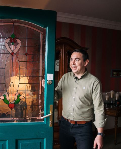 Een man staat bij een open glas-in-looddeur en lacht hartelijk. Hij draagt een lichtgroen shirt en een donkere broek. De deur heeft een rood en groen bloemenpatroon. Het interieur toont een warm verlichte woonkamer met gestreept behang, meubels en brochures op een tafel. | Echt Ierland Vakanties