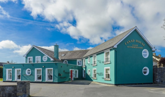 Fanad House is een groenblauw geschilderd gastenverblijf van twee verdiepingen in Kilkenny met witte sierranden en ronde ramen. Het heeft balkons en een schuin dak en staat onder een gedeeltelijk bewolkte hemel, met een stenen muur en oprijlaan aan de voorkant. | Echt Ierland Vakanties