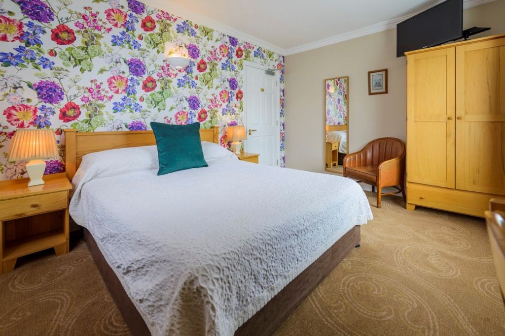 Een gezellige slaapkamer in Kilkenny guesthouse met een tweepersoonsbed bedekt met een witte sprei en een groen kussen. Kleurrijk bloemenbehang siert de muur en het houten meubilair bestaat uit nachtkastjes, een kledingkast, een bureau en een stoel. | Echt Ierland Vakanties