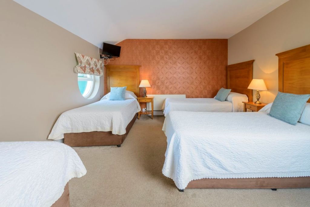 Een gastenkamer in Fanad House in Kilkenny met drie eenpersoonsbedden met wit beddengoed en blauwe kussens. Twee houten hoofdeinden rusten tegen een oranje accentmuur, plus een rond raam, lamp, tv hoog aan de muur en beige vloerbedekking. | Echt Ierland Vakanties