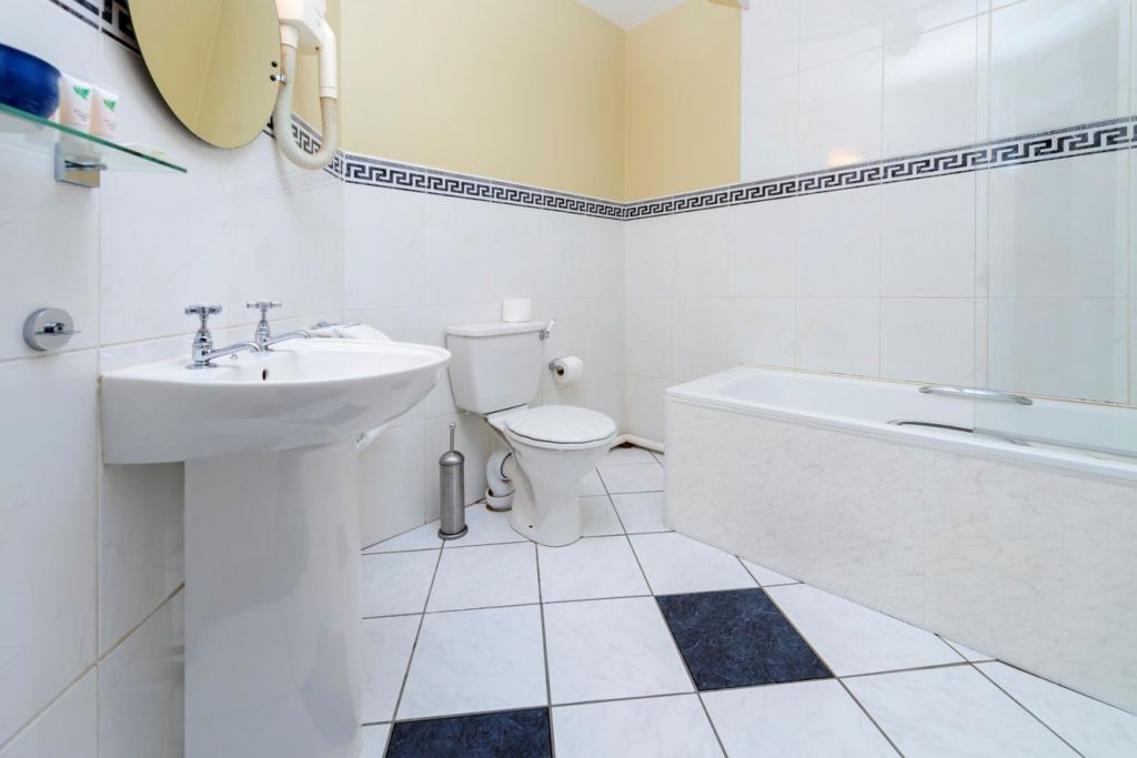 Een schone badkamer in het Fanad House guesthouse in Kilkenny heeft witte tegelvloeren met zwarte diamanten accenten, een wastafel op voet, een ronde spiegel aan de muur, een toilet met borstel ernaast en een bad met glazen paneel. De muren zijn betegeld en beige geschilderd. | Echt Ierland Vakanties