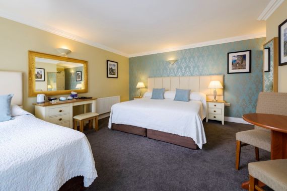 Een ruime kamer in het Fanad House in Kilkenny heeft twee bedden, nachtkastjes met lampen, ingelijste kunst op blauwe en gele muren, een grote spiegel, een tafel met stoelen en een theeservies. Zachte verlichting en vloerbedekking zorgen voor een warm welkom. | Echt Ierland Vakanties