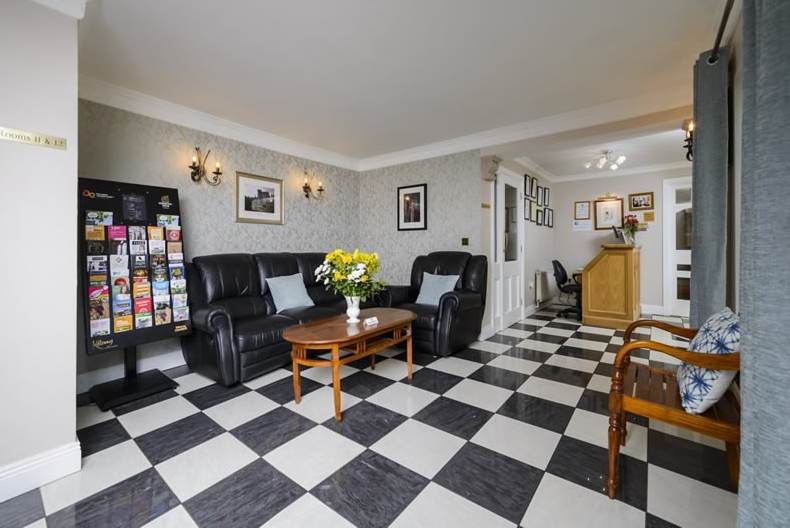 De lichte wachtkamer van het Fanad House guesthouse in Kilkenny heeft een zwart-wit geblokte vloer, zwarte banken, een houten salontafel met gele bloemen, een bankje, een folderstandaard, ingelijste foto's op muren met lichte patronen en een kleine houten receptiebalie. | Echt Ierland Vakanties