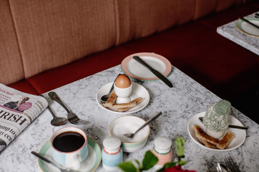 Een marmeren tafel herbergt een zachtgekookt ei in een eierdopje, toastreepjes en een kleine cactus; in de buurt staan koffie en een krant. Bestek en kruidenstrooiers zijn aanwezig. Een rode, gewatteerde bank weerspiegelt de gezellige sfeer van Ierland vakanties op de achtergrond. | Echt Ierland Vakanties