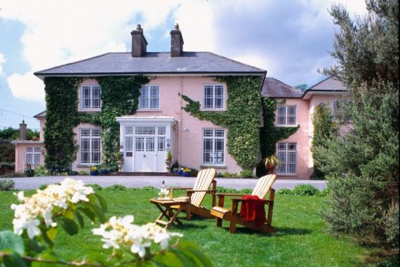 Een roze hotel met twee verdiepingen gelegen nabij Clifden Ierland, is versierd met klimop op delen van de gevel. Het heeft grote ramen en een veranda met witte pilaren. Op het weelderige groene gazon staan twee houten stoelen tegenover een kleine tafel met wijn, onder een gedeeltelijk bewolkte lucht omlijst door witte bloemen. | Echt Ierland Vakanties