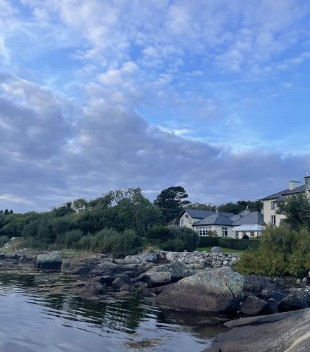 Een rustig kustlandschap met een rotsachtige kustlijn met kalm water op de voorgrond. Rechts is een groot huis met meerdere schoorstenen gedeeltelijk verduisterd door weelderig groen. De lucht erboven is uitgestrekt, gevuld met verspreide wolken die worden verlicht door zacht zonlicht. | Echt Ierland Vakanties