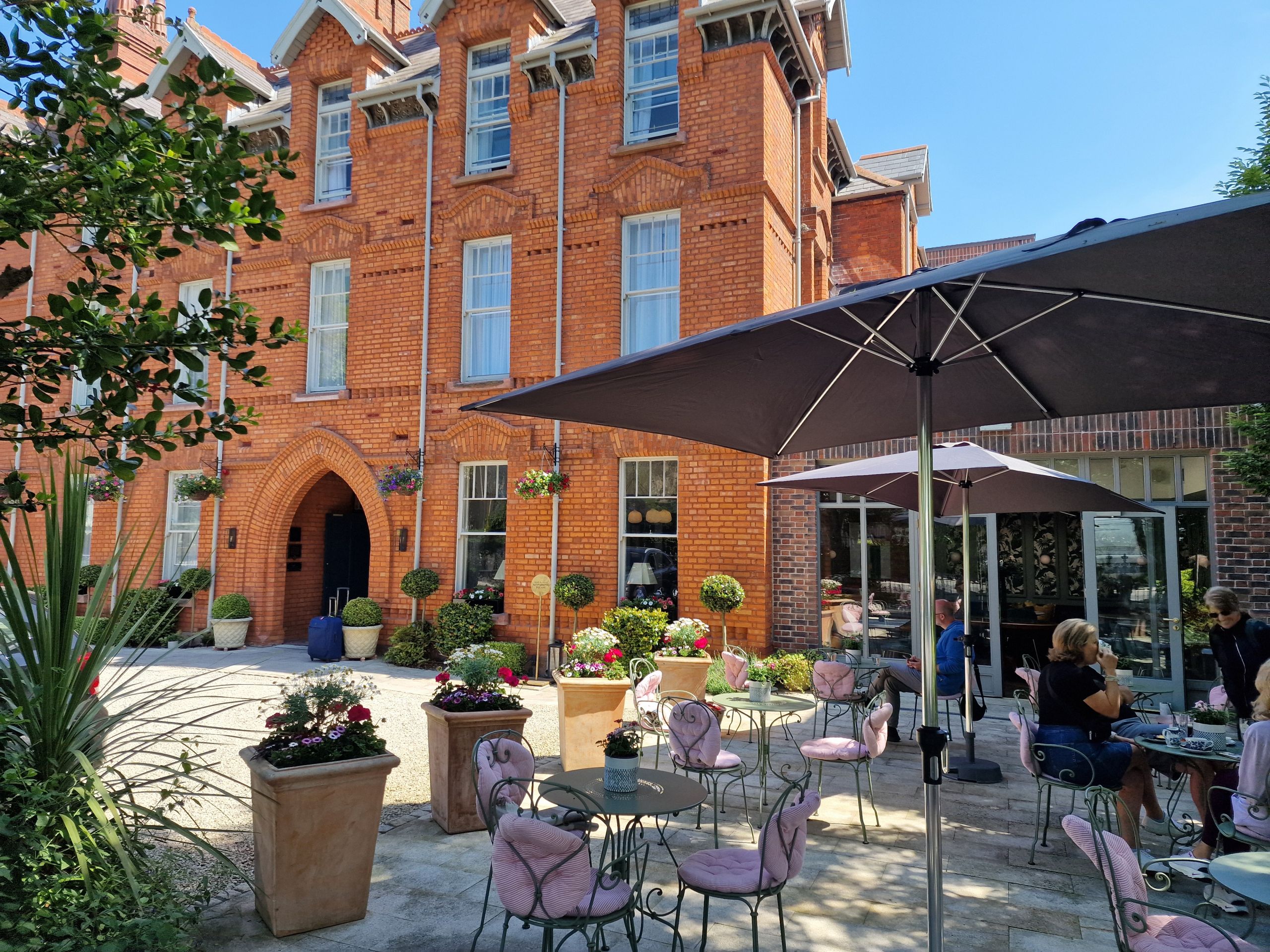 Het terras van een historisch bakstenen gebouw met een gewelfde ingang. Tafels en stoelen met roze kussens staan rond grote potplanten. Mensen zitten onder grote parasols en genieten van het zonnige weer. Het groen omringt het gebied en draagt bij aan de rustige sfeer. | Echt Ierland Vakanties