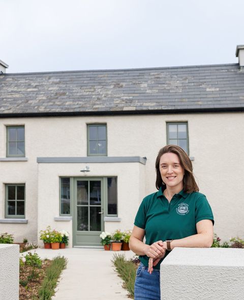 Een vrouw met lichtbruin haar, gekleed in een groene polo en spijkerbroek, staat glimlachend voor een crèmekleurig huis met grijze kozijnen, een leien dak en bloempotten bij de ingang. De tuin is netjes en er leidt een pad naar de deur. | Echt Ierland Vakanties