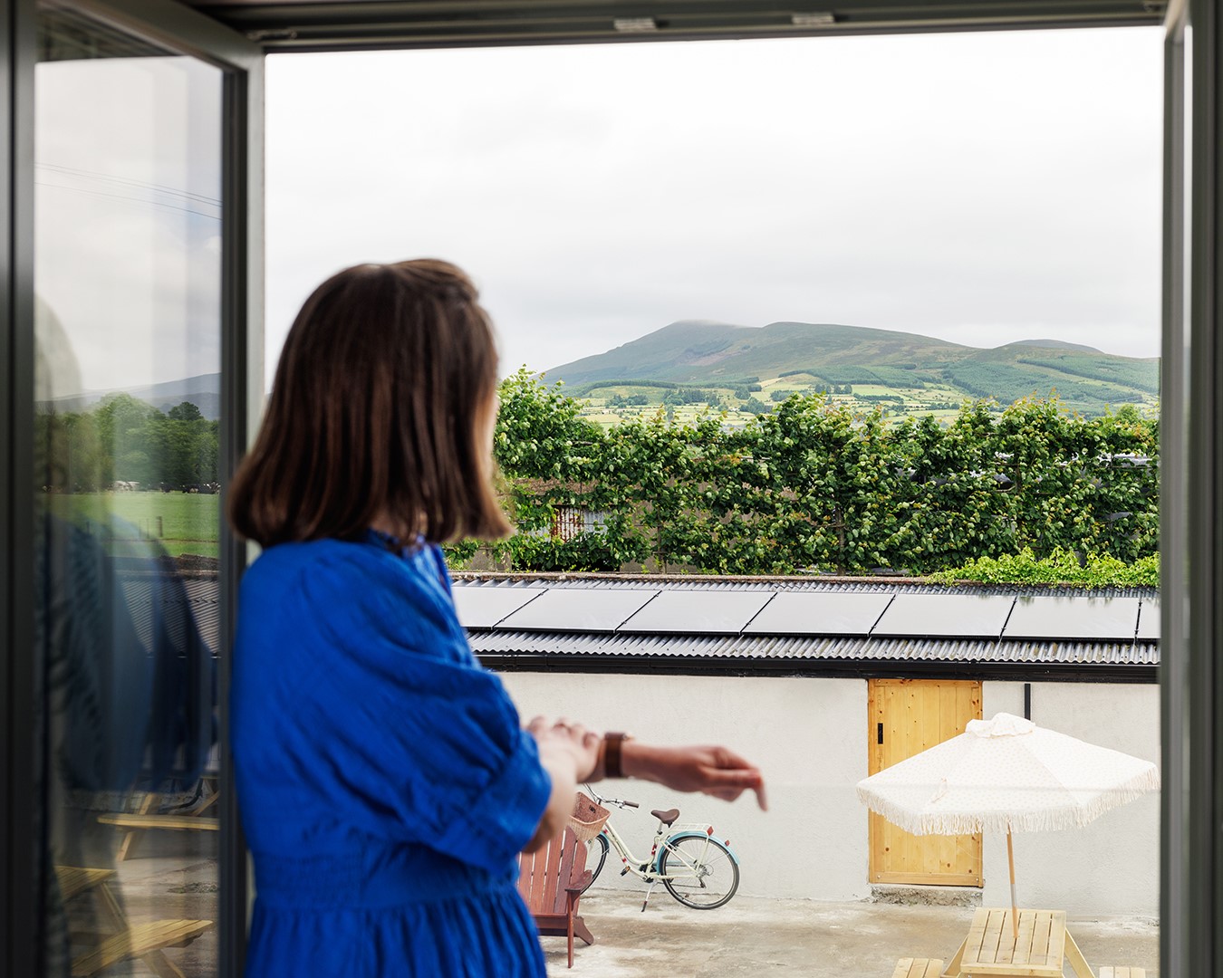 Een vrouw in een felblauwe jurk staat bij een open deur en kijkt naar buiten, naar een groen landschap met heuvels. Op de voorgrond staan een muur, een fiets, een parasol, een picknicktafel en zonnepanelen op het dak. Het tafereel oogt kalm en bewolkt. | Echt Ierland Vakanties