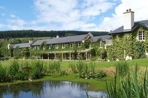 Groot landhuis met met klimop begroeide muren en hoge schoorstenen, te midden van weelderige groene gazons en tuinen. Een vijver met riet op de voorgrond, terwijl beboste heuvels en een gedeeltelijk bewolkte blauwe lucht de achtergrond vullen. Het tafereel is vredig en uitnodigend. | Echt Ierland Vakanties
