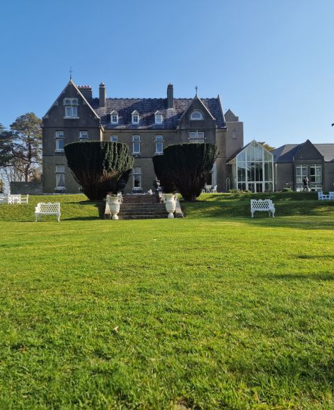 Een groot grijs stenen landhuis met dakkapellen staat achter een keurig onderhouden groen gazon. Een trap, geflankeerd door snoeiheggen, leidt naar de ingang. Witte banken en zitjes staan verspreid over het terrein, en hoge bomen omlijsten de helderblauwe lucht. | Echt Ierland Vakanties