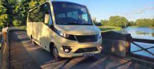 Een crèmekleurig minibusje met Marlowe op de voorruit staat geparkeerd op een smalle brug naast een kalme rivier, die de charme van een busrondreis Ierland vastlegt te midden van groene bomen en gras onder een strakblauwe hemel in deze vredige landelijke omgeving. | Echt Ierland Vakanties