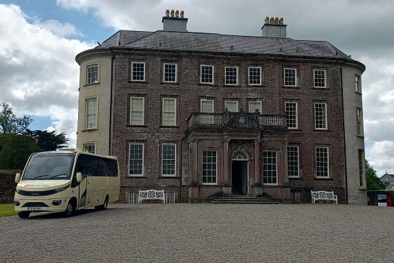 Een groot Georgisch herenhuis van drie verdiepingen met bruinrode bakstenen en witte sierranden staat onder een bewolkte hemel. Een crèmekleurige minibus, onderdeel van een busrondreis Ierland, staat geparkeerd op de oprit met grind. Bomen en een stenen muur omringen het statige gebouw. | Echt Ierland Vakanties