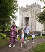 Een gezin van vier, met twee jonge kinderen, wandelt over een pad vlakbij het middeleeuws Bunratty Castle in county Clare. De moeder houdt de hand van haar dochter en een telefoon vast, terwijl de vader volgt met hun zoon. Weelderige bomen en gras omringen hen onder een bewolkte hemel. | Echt Ierland Vakanties