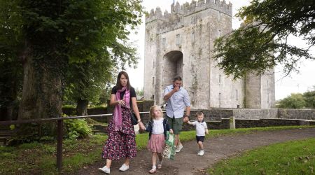 Een gezin van vier, met twee jonge kinderen, wandelt over een pad vlakbij het middeleeuws Bunratty Castle in county Clare. De moeder houdt de hand van haar dochter en een telefoon vast, terwijl de vader volgt met hun zoon. Weelderige bomen en gras omringen hen onder een bewolkte hemel. | Echt Ierland Vakanties