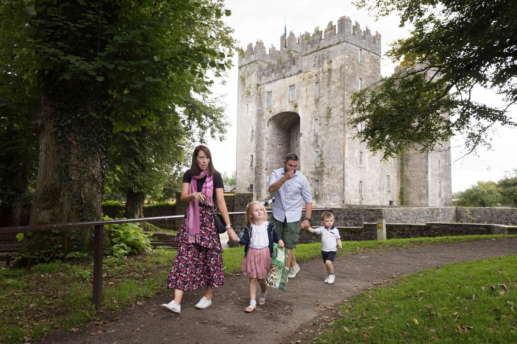 Een gezin van vier, met twee jonge kinderen, wandelt over een pad vlakbij het middeleeuws Bunratty Castle in county Clare. De moeder houdt de hand van haar dochter en een telefoon vast, terwijl de vader volgt met hun zoon. Weelderige bomen en gras omringen hen onder een bewolkte hemel. | Echt Ierland Vakanties