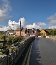 Een bochtige weg in het charmante vissersdorpje Doolin leidt langs kleurrijke huizen - oranje, roze en geel - tegen een bewolkte blauwe lucht. Een stenen muur omzoomt de Wild Atlantic Way, met gras begroeide heuvels en geparkeerde auto's die het schilderachtige landelijke landschap completeren. | Echt Ierland Vakanties
