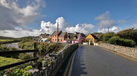 Een bochtige weg in het charmante vissersdorpje Doolin leidt langs kleurrijke huizen - oranje, roze en geel - tegen een bewolkte blauwe lucht. Een stenen muur omzoomt de Wild Atlantic Way, met gras begroeide heuvels en geparkeerde auto's die het schilderachtige landelijke landschap completeren. | Echt Ierland Vakanties