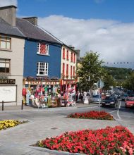 Een levendige straat in Westport aan de westkust van Ierland wordt gekenmerkt door kleurrijke winkelpuien zoals die van Flaherty en Thomas Moran, levendige bloemperken en wapperende vlaggen. Auto's en loofbomen omzomen de weg onder een halfbewolkte Ierse hemel. | Echt Ierland Vakanties